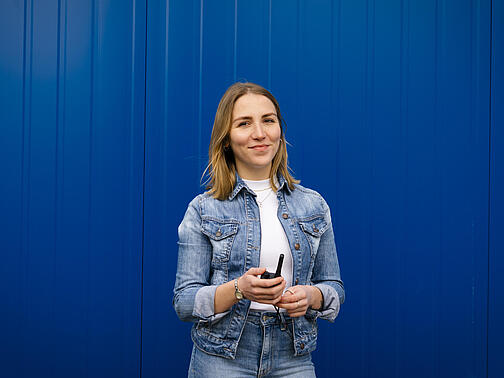 Die Mitarbeiterin steht vor einer blauen Containerwand mit einem Walkie-Talkie in der Hand.