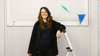 Kira steht vor einer weißen Containerwand und lehnt an einer Leiter.