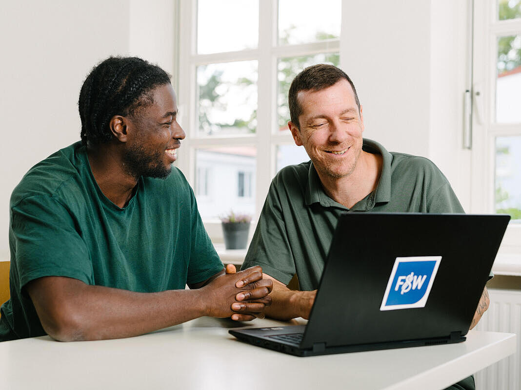 Zwei Männer sitzen in hellem Raum vor einem Laptop