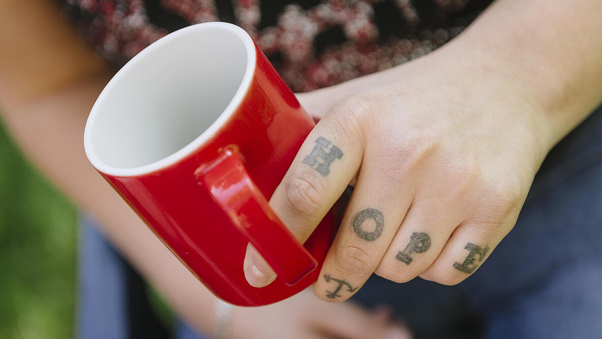 Eine Hand hält eine rote Tasse, auf den Fingern sind die Buchstaben "HOPE" tättowiert.