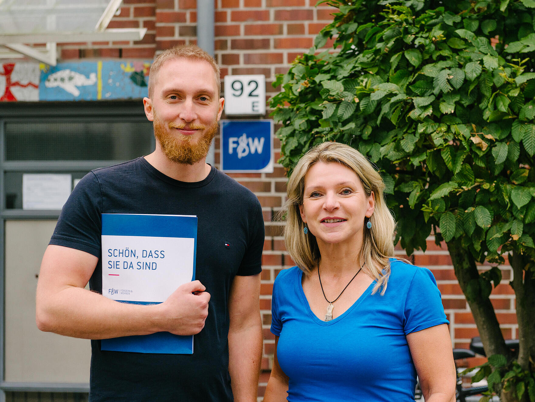 Zwei Mitarbeitende nebeneinander vor einem Haus